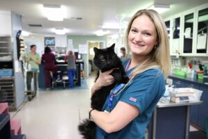 A blond smiling vet tech holds a black cat with one eye.