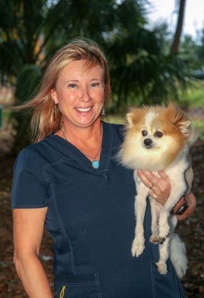 Dr. Helga Bleyaert, wearing navy blue scrubs, smiles while holding a fluffy, light-colored Pomeranian outdoors with trees and greenery in the background.