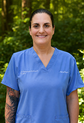 Dr. Cassandra Ruthrauff smiles outside in a wooded area. She is wearing light blue scrubs.