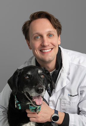 Dr. Jordan Yesesky smiles for a professional photo with his salt-and-pepper lab mix. He is wearing a black shirt and his BluePearl whitecoat.