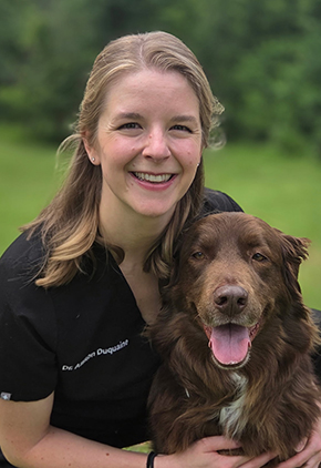 Dr. Allison Duquaine smiles outside while kneeling next to her brown dog. She is wearing black scrubs.