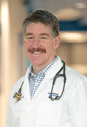 Dr. Kristopher Sharpe smiling in the lobby of the Grand Rapids hospital. He has on his BluePearl white coat, a blue and white checkered button-up, with a stethoscope around his neck.