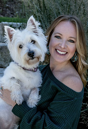 Dr. Aubrey Hnatusko smiles outside while holding her West Highland White Terrier. She is wearing a forest green off-the-shoulder sweater.