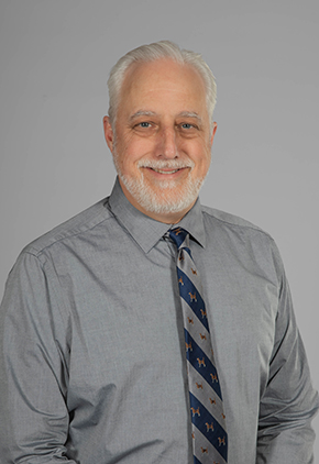 Dr. Lawrence Bagley smiles for a professional studio headshot. He is wearing a grey dress shirt and a navy and grey tie.
