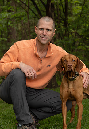 Dr. Michael Doornink smiles outside while kneeling next to his yellow Labrador. He is wearing grey dress pants and an orange quarter zip.