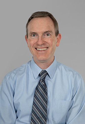 Dr. Michael Finn smiles for a professional headshot in front of a grey background. He is wearing a light blue dress shirt and a blue and grey striped tie.