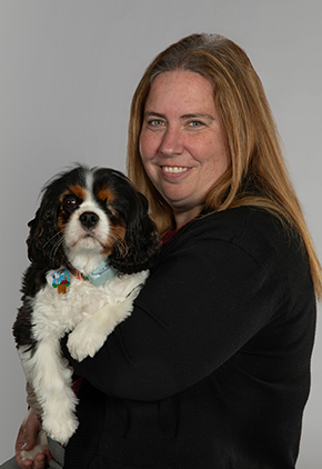 Dr. Kara Gornik smiles for a professional studio photo in front of a grey background while holding her black and white Saint Bernard mix in her arms. She is wearing a black blouse.