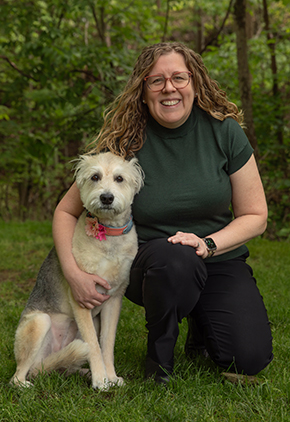 Dr. Rachel Keller Vaughn smiles outside in a lush wooded area. She is kneeling next to her dog and wearing black pants and a green blouse.