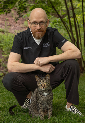 Dr. Edward Mackillop poses outside in a lush, open area, kneeling in front of his tabby cat. He is wearing black scrubs and black & white checkered shoes.