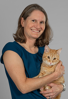 Dr. Katrin Saile smiles for a professional headshot in front of a grey studio background. She is wearing a teal blouse and holding her orange cat.