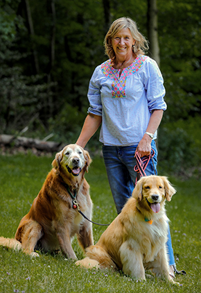 Dr. Lisa Sepesy smiles outside in a lush, wooded area. She has the leash of her two golden retrievers in each hand. She is wearing a blue blouse and blue jeans.