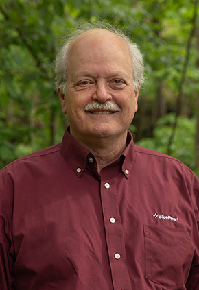 Dr. Anthony Pardo smiles for a professional headshot outside in front of lush woods. He is wearing a maroon button-up.
