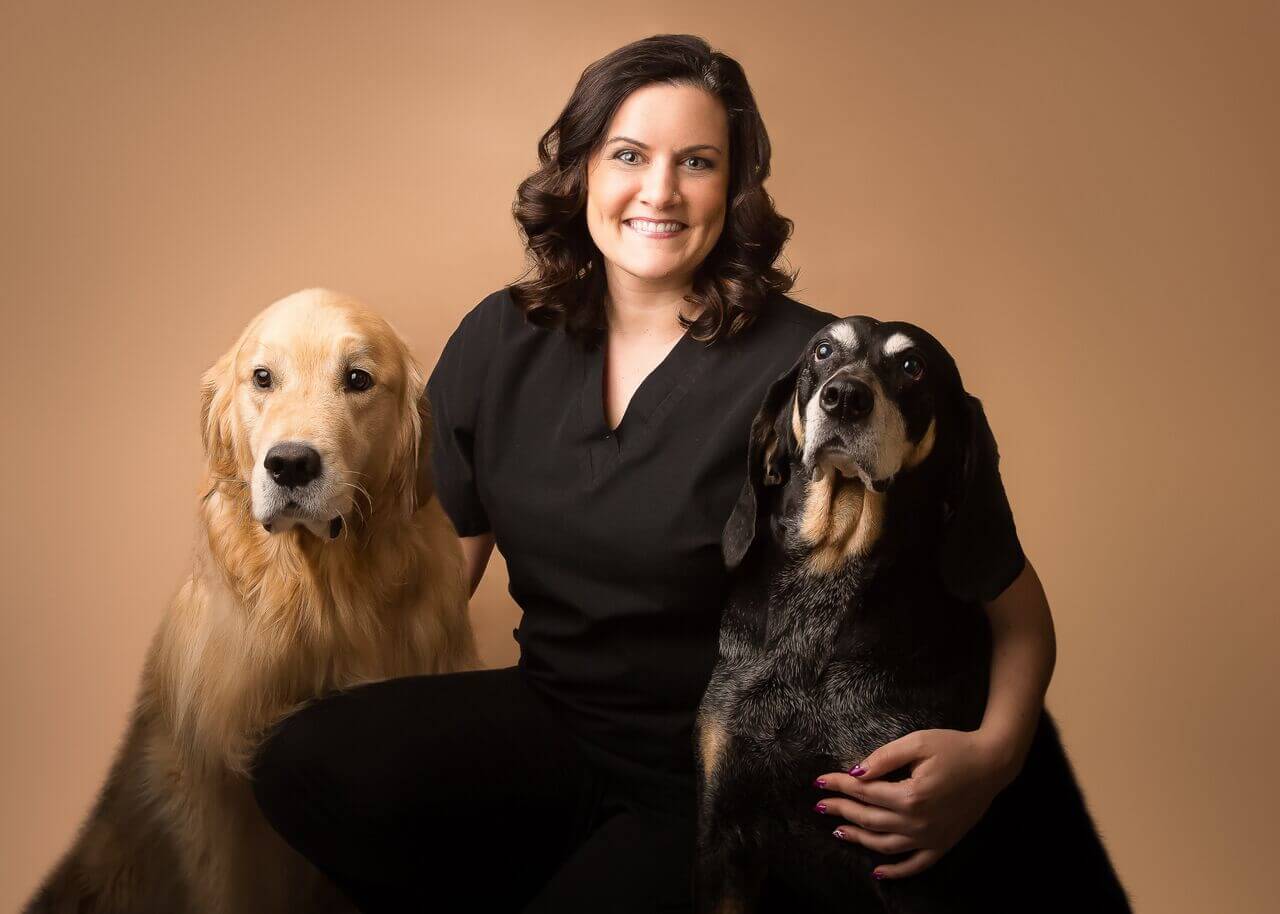A smiling brunette poses with her arms around two dogs.