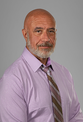 Dr. Samuel Durkan smiles for a professional headshot in front of a grey background. He is wearing a powder pink dress shirt and pink and grey striped tie.