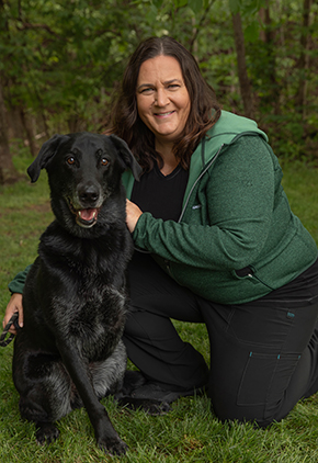 Dr. Michelle Fulks smiles outside while kneeling next to her black Labrador. She is wearing black scrubs and a heather green full-zip jacket.