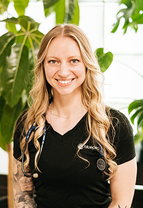 Dr. Alexandria Baltes smiles for a professional headshot in front of a window and a large green plant. She is wearing a black BluePearl v-neck and has her stethoscope around her neck.