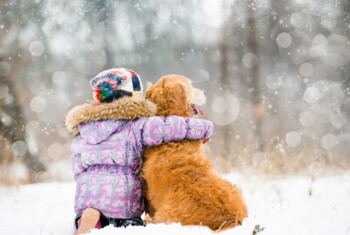 A girl in a purple coat hugs a Golden Retriever next to her while sitting in the snow.