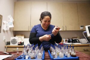 A smiling tech prepares a vaccine from numerous glass bottles on a table.