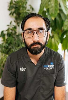 Dr. Amanpreet Grewal sitting with a soft smile in front of a large, green, leafy plant. He has on glasses and a dark grey set of BluePearl scrubs.