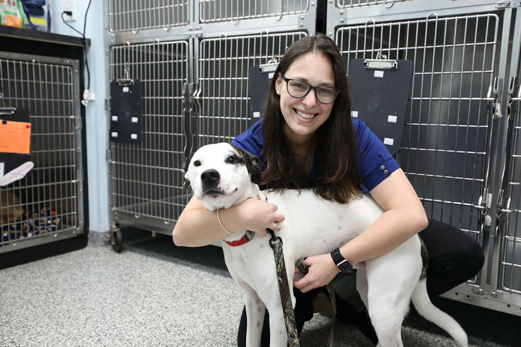 A technician hugs a white dog close to her with kennels in the back.