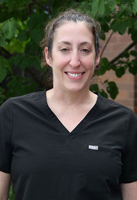 Dr. Alexandra Kalamaras smiles outside in front of a green, leafy tree. She is wearing black scrubs.