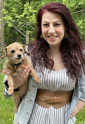 Dr. Laura Scoda smiles outside while holding her terrier mix, Primrose. She is wearing a white dress that features thin black stripes. She has on a grey jacket and a wide brown belt around her waist.
