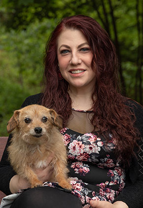 Dr. Laura Scoda smiles outside in a lush, wooded area. She is wearing a black floral top and holding her terrier mix.
