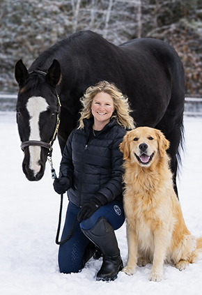 Dr. Stephanie Brock smiles outside in a snowy field. She has her black horse to her right and a smiling golden retriever to her left.