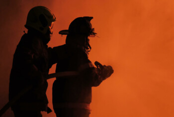 Two fire fighters walk through red hued smoke.