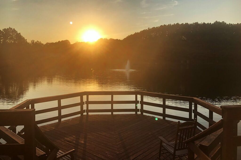 A deck extends over a lake while the sun rises above the tree line.