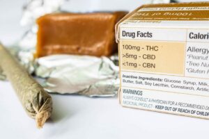 A hand-rolled cigarette, box of medicine containing 100mg - THC, >5mg - CBD, <1mg - CBN; and a brick of tan solids rest on a table.