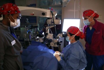 An ophthalmologist in a blue gown performs surgery using a microscope.