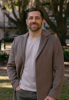 Dr. Marcus Bradbury smiles outside on a fall day. He is wearing dark grey jeans, a light grey t-shirt and a dark taupe peacoat.