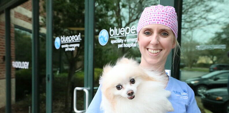 A BluePearl associate holds a dog while smiling in front of a BluePearl Pet Hospital.