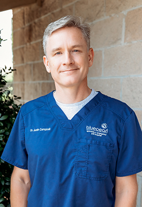 Dr. Justin Campbell smiling outside in front of a brick building. He is wearing dark blue BluePearl scrubs.