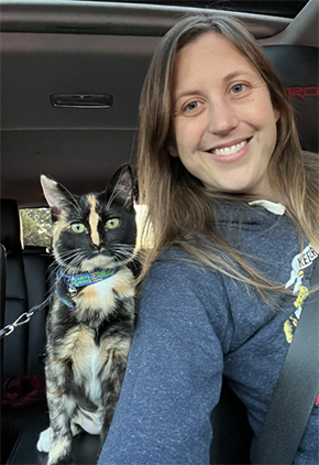 Dr. Becky Skover smiles from the front seat of her parked car while taking a selfie with her black and white cat. She is wearing a seatbelt, and the cat is secured with a leash for safety.