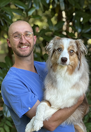 Dr. Jackson Sanders smiles outside while holding up his white and tan border collie while she crosses her legs. He is wearing light blue scrubs.