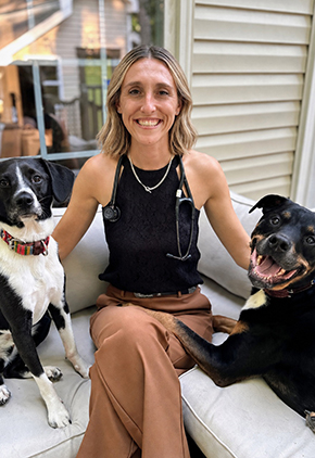 Dr. Courtney Bounds smiles outside on her deck couch with a black dog on either side of her. She is wearing a black shirt, tan slacks and has a stethoscope around her neck.