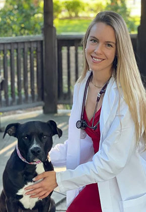 Dr. Karina Payerhin smiles outside while kneeling next to her black pit bull. She is wearing a red dress, her white coat and has a stethoscope around her neck.
