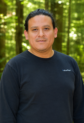Dr. Rothman Reyes softly smiles outside in a wooded area. He is wearing a navy BluePearl long-sleeve shirt.