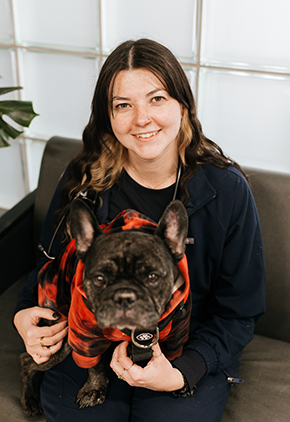 Dr. Emily Davis smiles for a photo with her black bulldog. She is wearing a black sweater and her dog has on a red jacket.