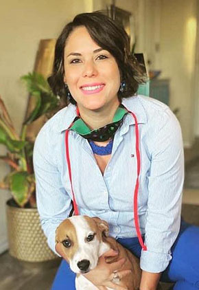 Dr. Yalisa Figueroa smiles while kneeling and holding her hound mix. She is wearing blue scrubs and a light BluePearl jacket.
