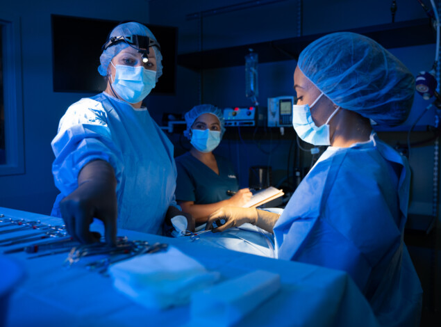 A veterinarian and two technicians in scrubs and protective gear prepare surgical instruments.