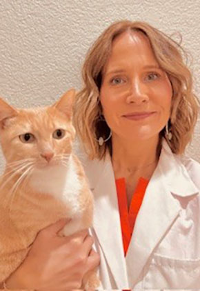 Dr. Michelle Hazlewood smiles in front of a white wall while holding an orange cat. She is wearing a coral top and her whitecoat.