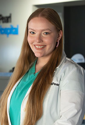 Dr. Natalie Timmons is an emergency veterinarian. She has long red hair and is wearing her teal scrubs and white coat. She's standing in the lobby of BluePearl and smiling.