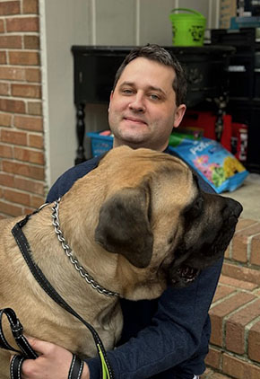Dr. Paul Rossman sitting outside a garage. He has a smile on his face while hugging his Boerboel .