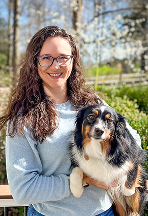 Dr. Sarah Frankel-Freer smiles outside on a spring day. She is wearing a powder blue sweater and holding one of her Australian shepherds in her arms.