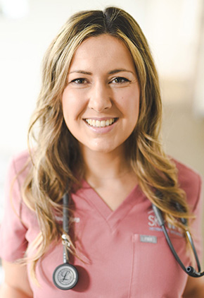 Dr. Natalie Allers, wearing a rose-colored scrub top and a stethoscope around her neck, smiles warmly at the camera in a well-lit indoor setting.