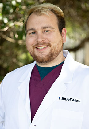 Dr. Michael Karchner smiling outside in front of a tree. He is wearing maroon scrubs, a dark grey tee and his BluePearl whitecoat.