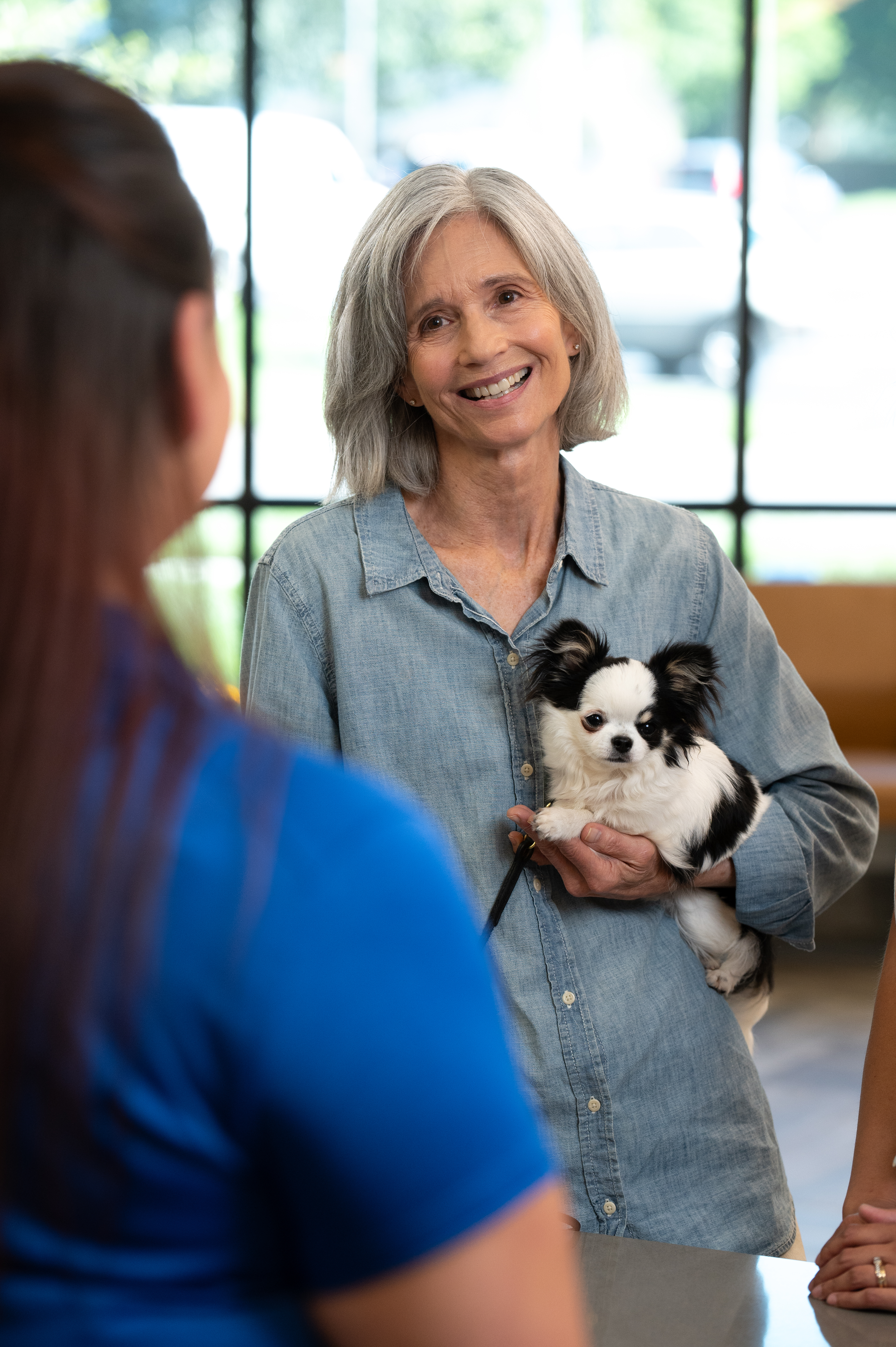 A woman smiles at another woman while holding a small dog in her arms.
